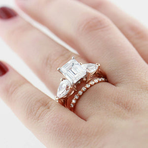 Large three stone engagement ring with emerald cut and pear cut diamond hybrid simulants set in 14k rose gold stacked next to diamond accented band worn on hand