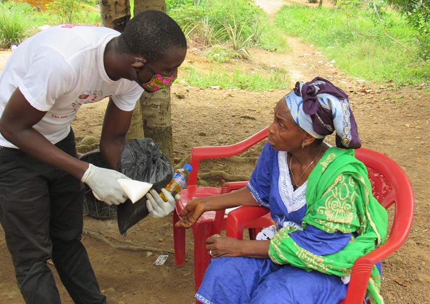 A mask-clad person with The Greener Diamond partnership COVID urgent relief program in Liberia assists an elderly seated woman