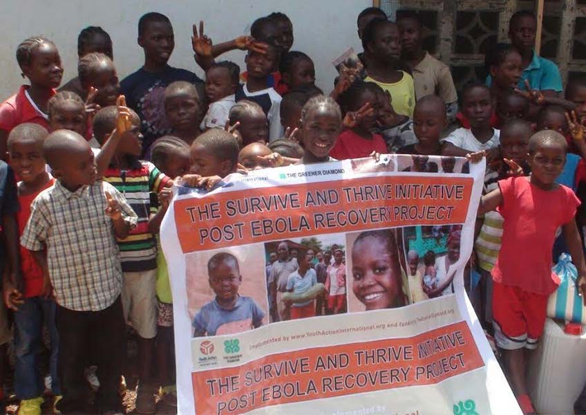A group of children hold up a banner featuring The Greener Diamond logo and promoting the Survive and Thrive Initiative Post Ebola Recovery Project as part of the EBOLA PROJECT in LIBERIA