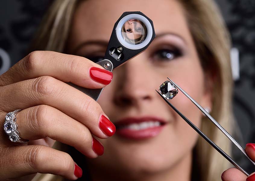 An image of MiaDonna founder Anna-Mieke Anderson, wearing a white gold three stone engagement ring, using a loupe to inspect a large cushion-cut lab created diamond held with diamond tweezers.