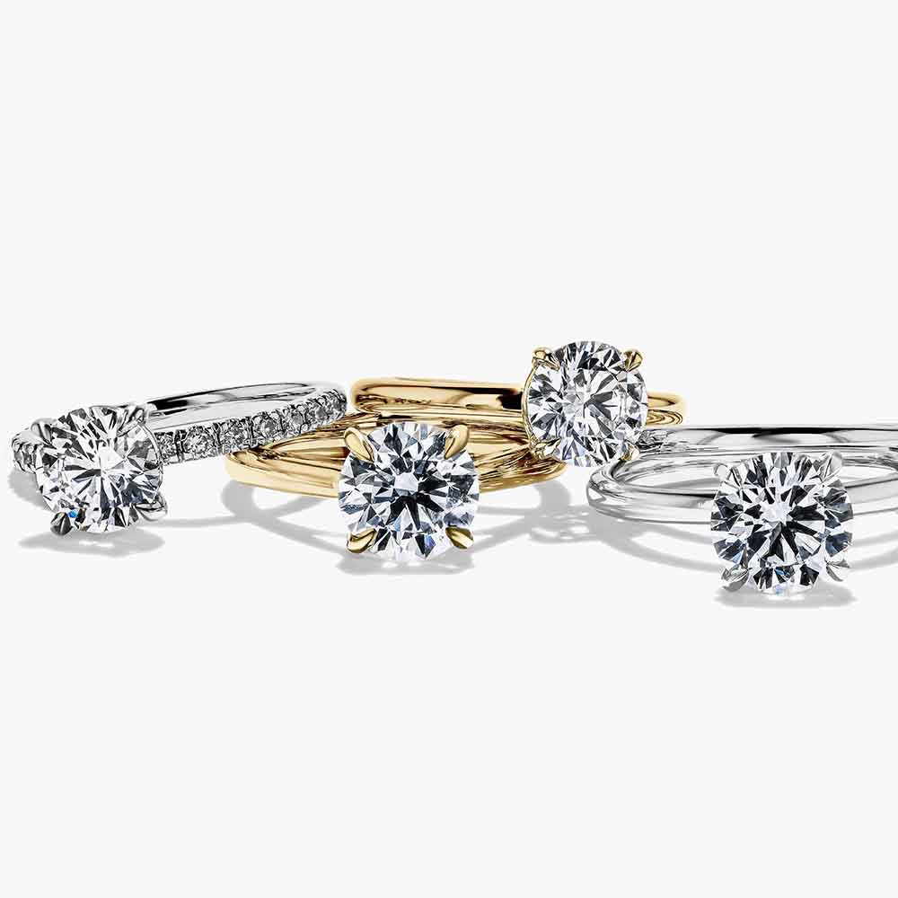A collection of MiaDonna engagement rings in various colors, including gold, silver, rose gold, and platinum, each featuring a round cut lab created diamond center stone.
