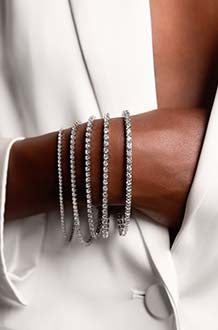 A stylish woman wearing a white blazer demonstrates five different stone size options for MiaDonna's conflict-free, lab-created diamond tennis bracelets by displaying them stacked along her wrist..