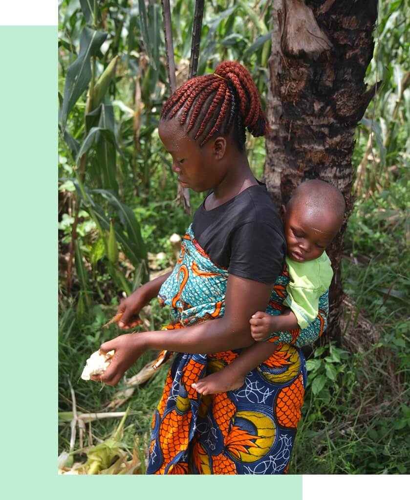 At a The Greener Diamond location in Liberia, a woman carries a baby in a sling while standing among greenery.