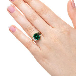 Three stone engagement ring with 2ct emerald cut lab created emerald center between baguette cut lab diamonds set in 14k yellow gold worn on hand