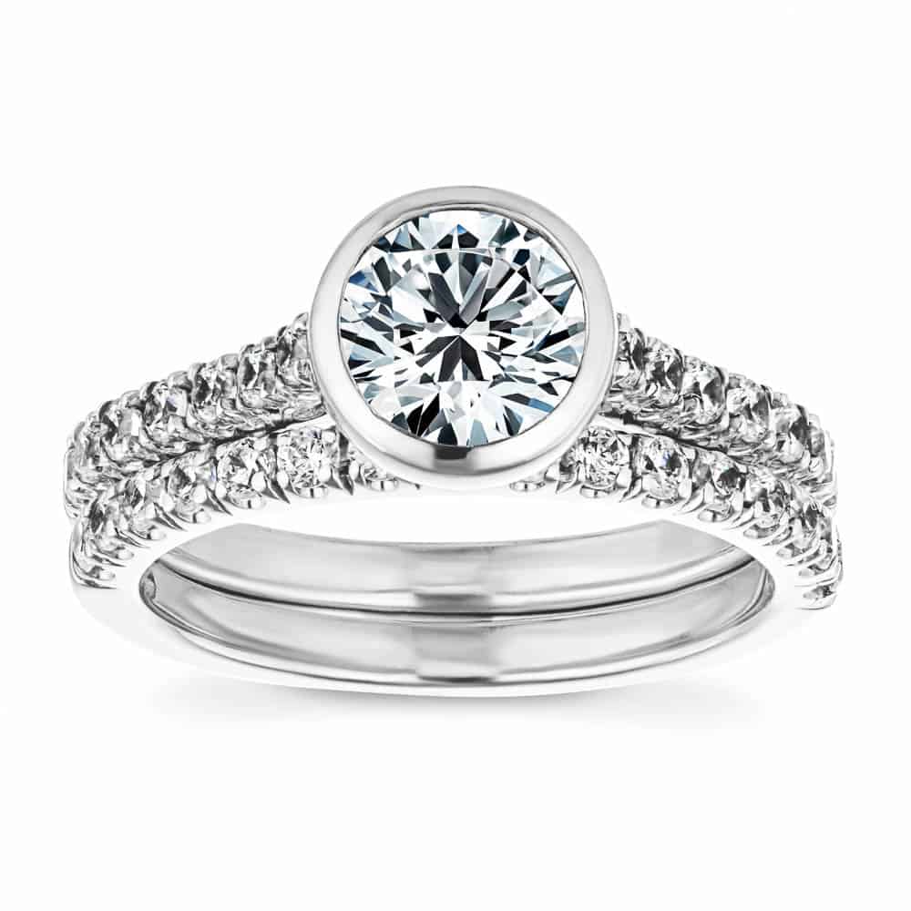 Shown with a bezel set 1.0ct Round cut Lab-Grown Diamond with accenting stones on the band in recycled 14K white gold with matching wedding band, can be purchased as a set for a discounted price 